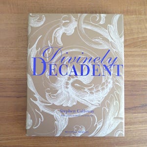 May include: A hardcover book titled "Divinely Decadent" by Stephen Calloway. The book cover is a light tan color with white floral designs and blue text. The book is lying on a wooden surface.