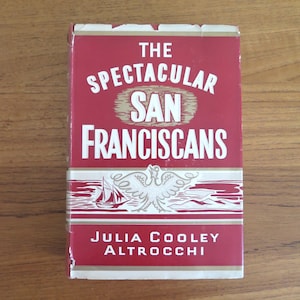 May include: A hardcover book titled The Spectacular San Franciscans by Julia Cooley Altrocchi. The book cover is deep red with white text and features a white graphic illustration of a bird and a sailing ship centered between the title and author.