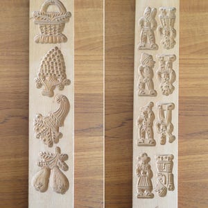 May include: Two vintage wooden cookie molds. One mold features a basket, grapes, a cornucopia, and pears. The other mold has figures of people and buildings. Both molds are made of light-colored wood.