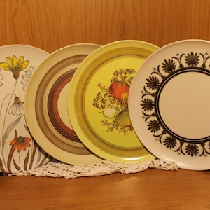May include: Four vintage ceramic plates with different floral designs. The plates are white with brown, yellow, and green accents. The designs include a floral pattern with a daisy, a floral pattern with a fruit, a concentric circle pattern, and a floral pattern with a border.