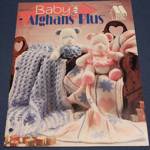 May include: Crochet Baby Afghans Plus pattern book featuring two crocheted teddy bears, one blue and one pink, sitting on a wooden crib with crocheted blankets. The book cover has the title "Crochet Baby Afghans Plus" and the logo "Annie's Attic".
