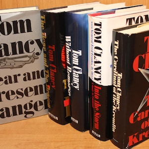 Może przedstawiać: Kolekcja powieści Toma Clancy'ego, w tym "Clear and Present Danger" i "The Cardinal of the Kremlin". Książki mają różne kolory grzbietów i okładek, z tytułami pogrubioną czcionką. Okładka "Clear and Present Danger" przedstawia helikopter.