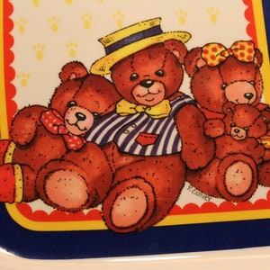 Könnte beinhalten: Eine weiße Keramik-Schale mit blauem Rand und einem roten, gelben und weißen gewellten Rand. Die Schale zeigt ein Design mit vier braunen Teddybären mit einem blau-weiß gestreiften Hemd, einer roten Schleife und einer roten Schleife mit einer gelben Blume. Der Text "F. Carney" ist in der rechten unteren Ecke der Schale aufgedruckt.