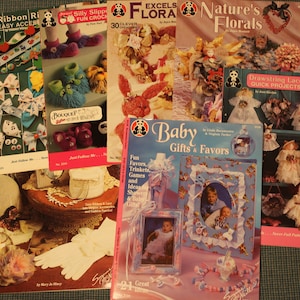 Könnte beinhalten: Eine Sammlung von Bastelbüchern mit verschiedenen Projekten, darunter Bänderarbeiten, Blumenarrangements, Babygeschenke und Kordelspitzenprojekte. Die Bücher haben farbenfrohe Cover mit Illustrationen und Text.