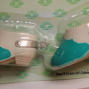 May include: Pair of doll shoes in a plastic package. The shoes are two-tone, with a cream-colored upper and a turquoise toe. Each shoe has a silver-colored strap and a star-shaped spur. The package reads "Shoes fit 14" Cabbage Patch Kids® Only!"