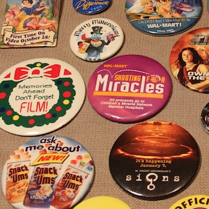 Puede incluir: Una colección de botones vintage con varios personajes y logotipos de películas y programas de televisión. Los botones son coloridos y presentan una variedad de diseños, incluyendo una corona roja y verde con las palabras "Memories Ahead Don't Forget FILM!" y un botón morado con las palabras "Shooting for Miracles" y el texto "All proceeds go to Children's Miracle Network Member Hospitals".