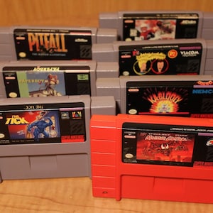 Könnte beinhalten: Eine Sammlung von acht Super Nintendo Entertainment System (SNES) Spielkassetten. Die Kassetten sind in einer Reihe angeordnet, wobei die Vorderseiten der Etiketten zum Betrachter zeigen. Die Kassetten sind alle in verschiedenen Farben gehalten, und die Etiketten zeigen verschiedene Figuren und Logos. Die Kassetten sind alle in gutem Zustand, ohne sichtbare Kratzer oder Beschädigungen.
