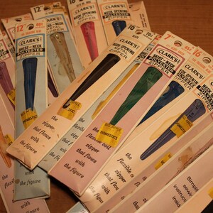 May include: A collection of vintage Clark's brand zippers in various colors, including blue, pink, and green. The packages display the zipper length in inches, with text that reads "Dress-Neck Polyester Zipper" and "Skirt-Neck Polyester Zipper".