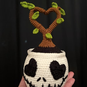 May include: A crocheted planter with a white pot, black heart-shaped eyes, and a stitched smile. A brown tree with a heart-shaped top and green leaves grows from the pot. A whimsical, handmade design, perfect for home decor.