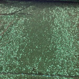 Op de afbeelding: Een rol smaragdgroene paillettenstof. De stof is bedekt met kleine, ronde pailletten die licht reflecteren en een glinsterend effect creëren. De pailletten zijn dicht op elkaar gepakt, waardoor de stof een luxueuze uitstraling heeft.