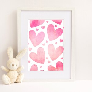 May include: A white frame containing a print with a pink watercolour heart pattern. The print features a repeating pattern of pink hearts of different sizes. The frame is sitting on a white surface next to a beige stuffed bunny.