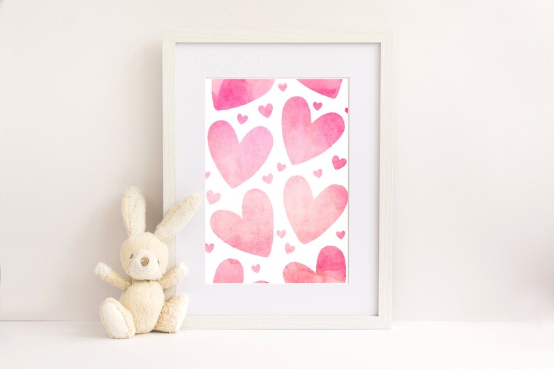 May include: A white frame containing a print with a pink watercolour heart pattern. The print features a repeating pattern of pink hearts of different sizes. The frame is sitting on a white surface next to a beige stuffed bunny.