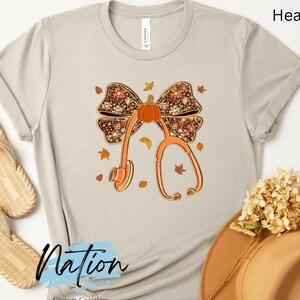 May include: A light beige t-shirt with a fall-themed graphic featuring a stethoscope, pumpkin, and bow. The design includes autumn leaves and the text "Heather Dust". The shirt is paired with woven sandals and a brown hat.