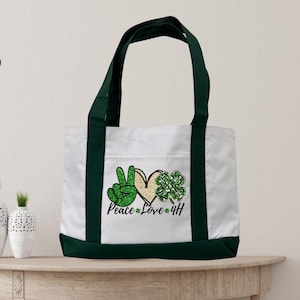 Puede incluir: Una bolsa de lona blanca con bordado verde y dorado de un signo de paz, un corazón y un trébol. La bolsa tiene asas verdes y el texto "Peace Love & HH" está bordado en la parte delantera.