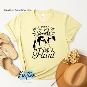 May include: A yellow t-shirt with the text "In a field of Sorrels be a Paint" and a black and white silhouette of a horse. The shirt is on a white background with a straw hat and sandals.