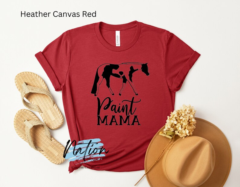 K&ouml;nnte beinhalten: Rotes T-Shirt aus Heather-Canvas mit einer schwarzen Silhouette eines Paint-Pferdes und eines Reiters. Der Text "Paint Mama" ist unter dem Bild gedruckt.