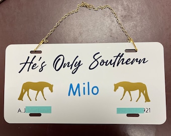 May include: White metal sign with a gold chain for hanging. The sign has the text "He's Only Southern" in blue cursive lettering, the name "Milo" in blue lettering, and two gold silhouettes of horses. The sign also has the text "A.J." and "921" in blue lettering.