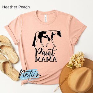 K&ouml;nnte beinhalten: Ein T-Shirt in der Farbe Heidepfirsich mit einer schwarzen und wei&szlig;en Silhouette eines Paint Horses und dem Text "Paint Mama".