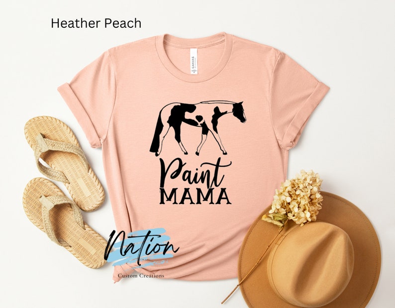 K&ouml;nnte beinhalten: Ein T-Shirt in der Farbe Heidepfirsich mit einer schwarzen und wei&szlig;en Silhouette eines Paint Horses und dem Text "Paint Mama".