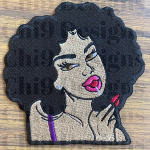 May include: Embroidered patch of a woman with an afro hairstyle, wearing a purple and beige top, and holding a lollipop. Her lips are pink and red, and she has brown eyes.