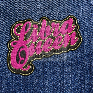 May include: A pink embroidered patch with the words "Libra Queen" in a cursive font. The patch has a black border with gold stitching and is set against a blue denim background.