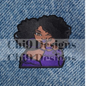 May include: Embroidered iron-on patch featuring a woman with brown skin, black hair, and purple clothing. She is winking and has a purple lip. The patch is on a blue denim background.