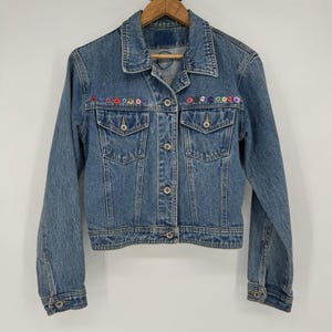 May include: A light blue denim jacket with a classic collar, button-down front, and two chest pockets. The jacket is adorned with colorful embroidered floral accents along the top. The jacket is hanging on a wooden hanger.