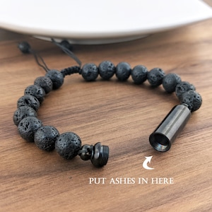 May include: Black lava rock beaded bracelet with a small metal cylinder attached. The cylinder has a screw-on top and the text "PUT ASHES IN HERE" is printed below the cylinder.