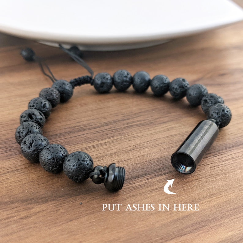 May include: Black lava rock beaded bracelet with a small metal cylinder attached. The cylinder has a screw-on top and the text "PUT ASHES IN HERE" is printed below the cylinder.