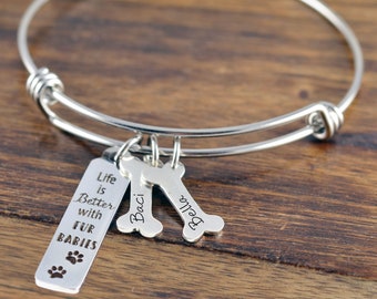 May include: Silver expandable bangle bracelet with three charms. The charms are a bone-shaped tag with the name "Baci" engraved on it, a bone-shaped tag with the name "Bella" engraved on it, and a rectangular tag with the text "Life is Better with Fur Babies" and a paw print.