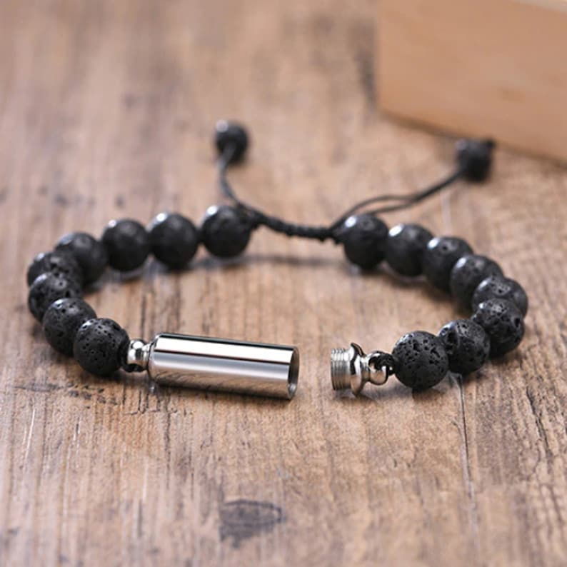 May include: A black lava rock beaded bracelet with a silver, cylindrical,  fillable charm. The charm is open and the bracelet is untied.