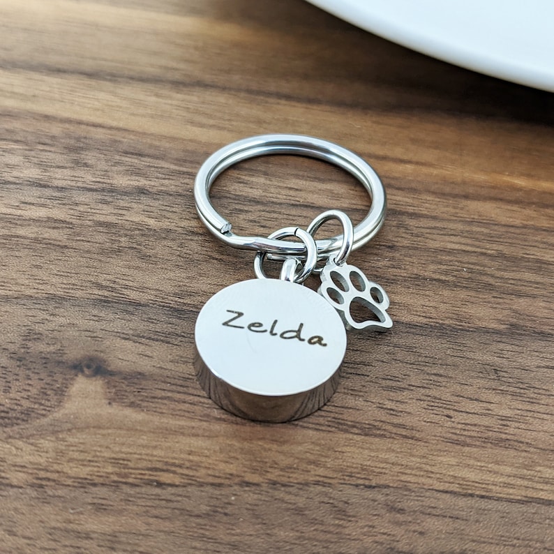 Puede incluir: Llavero de plata con un dije redondo grabado con el nombre "Zelda" y un peque&ntilde;o dije de huella de pata.