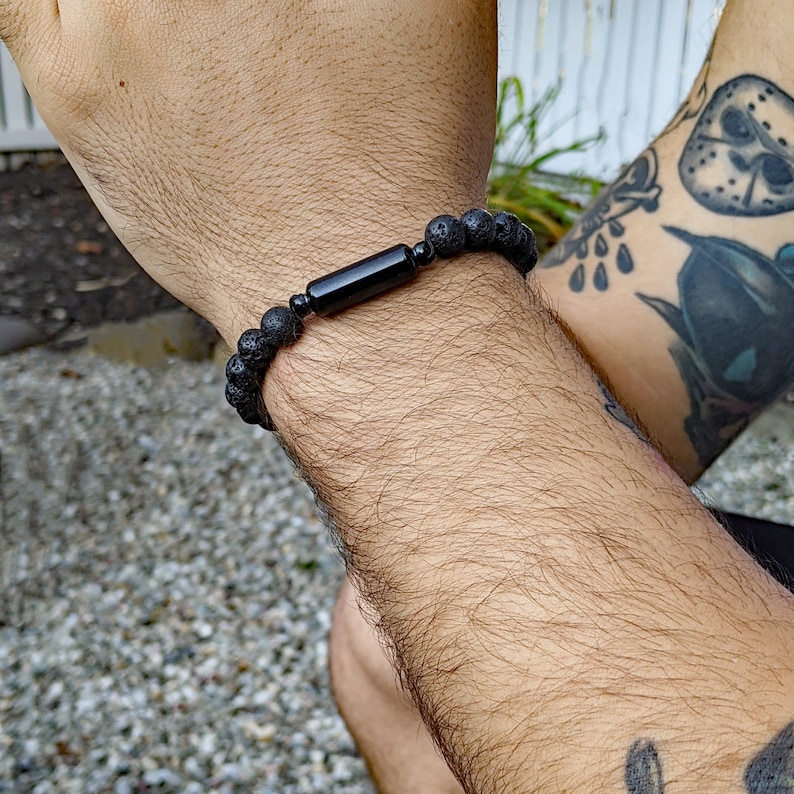 May include: A black lava rock bracelet with a black onyx bead. The bracelet is on a person's wrist.
