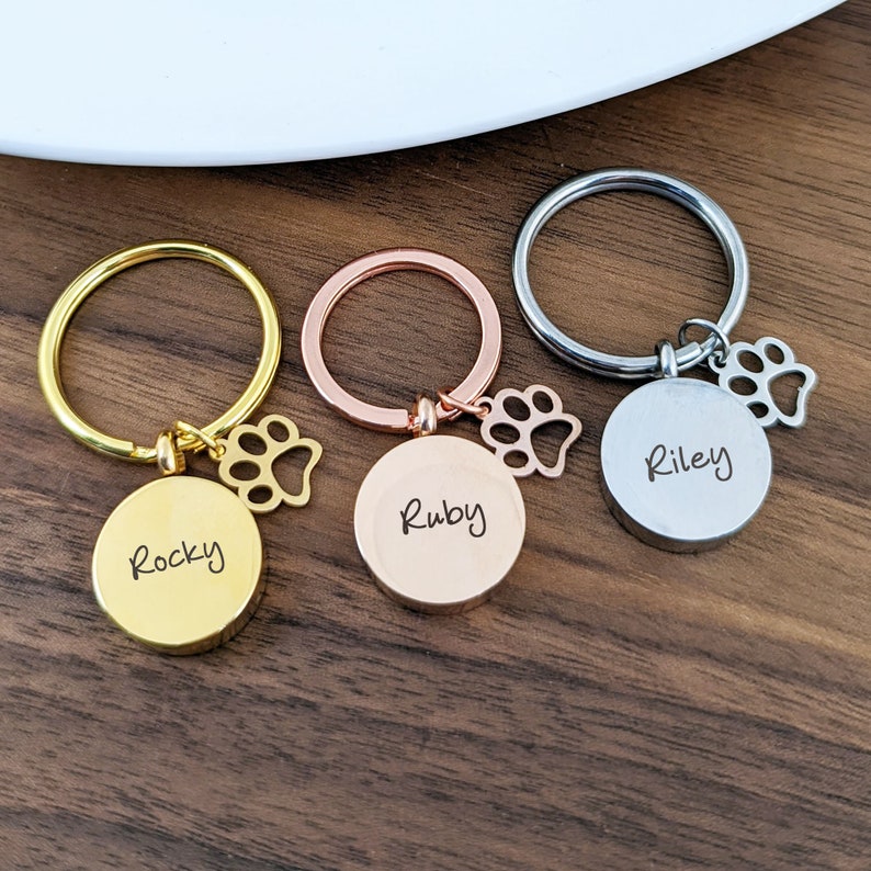 Puede incluir: Tres llaveros en oro, oro rosa y plata con dijes de huella de pata y nombres grabados "Rocky", "Ruby" y "Riley".