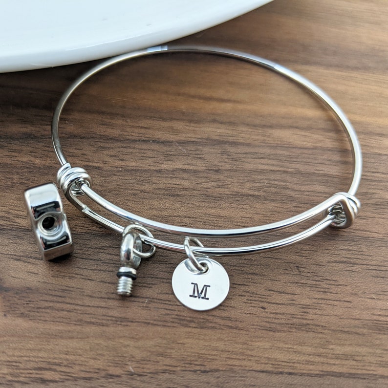 Pu&ograve; includere: Un braccialetto espandibile in argento con un piccolo ciondolo che dice "M" e un ciondolo a forma di vite.