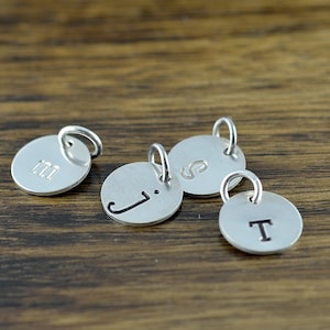 May include: Four silver charms with initial letters J, S, T, and M engraved on them. The charms are round and have a small loop at the top for attaching to a necklace or bracelet.