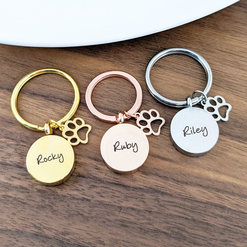 Puede incluir: Tres llaveros personalizados con dijes de huella de pata. Los llaveros son de oro, oro rosa y plata. Los nombres "Rocky", "Ruby" y "Riley" est&aacute;n grabados en los llaveros.