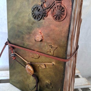 Op de afbeelding: Een handgemaakt notitieboek met een verweerde, getextureerde kaft in groen- en goudtinten. Een fiets en een vlinder zijn in reliëf op de kaft aangebracht. Het notitieboek is gebonden met een bruin leren koord en heeft verouderde papieren pagina's.