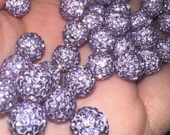 May include: A collection of sparkling, lavender-colored beads. Each bead is round and covered in small, glittering crystals, creating a textured, reflective surface. The beads are held in a hand, showcasing their size and detail. These beads are ideal for jewelry making.