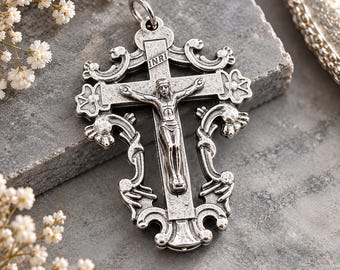 May include: A silver-toned metal crucifix pendant featuring a detailed figure of Jesus on the cross. The cross has ornate, swirling scrollwork edges and the letters INRI inscribed at the top. It includes a small metal loop for hanging.