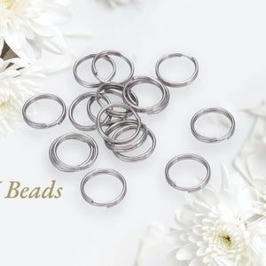 May include: A collection of silver-colored split key rings scattered on a white surface, with white flowers in the background. The key rings are made of metal and have a split design for easy attachment. The text "RJ Beads" is visible in the lower left corner.