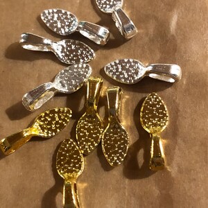 May include: A collection of silver and gold-colored spoon-shaped jewelry findings. The spoons have a textured bowl design and a loop for attaching to other jewelry components. The findings are scattered on a brown surface.