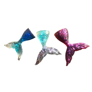 May include: Three glittery mermaid tail charms in blue, teal, and pink. The tails have a textured, scale-like appearance and are set against a white background. These decorative charms are ideal for crafting and jewelry making.
