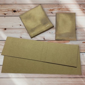 May include: Kraft paper notebooks and folded paper sheets. The notebooks are rectangular with a stapled binding. The paper sheets are folded with vertical score lines. The paper is a light brown color, and the background is a light brown wood grain.