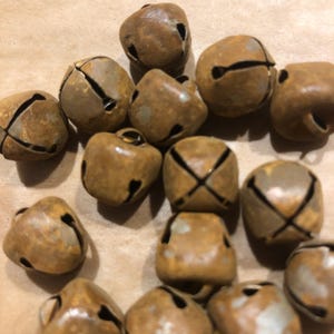 May include: A collection of small, weathered bronze-colored bells. Each bell has a slit and a small hole, with some featuring an 'X' design. The bells are arranged on a light brown surface.