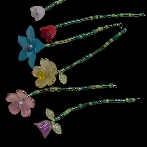 May include: Six floral hair accessories with translucent petals in white, blue, red, yellow, pink, and purple. Each flower is attached to a green beaded stem. The stems are adorned with various shades of green beads.
