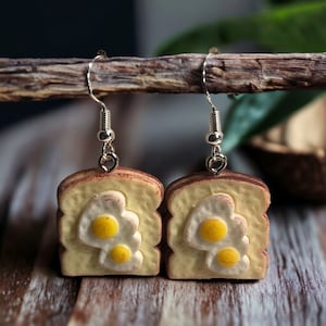 May include: A pair of dangle earrings featuring a miniature, detailed replica of a slice of toast with fried eggs. The earrings are made of polymer clay and have silver hooks.