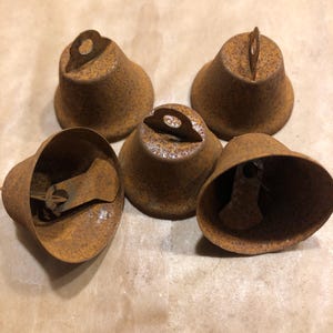May include: Five small, rusty brown metal bells with a bell shape. Each bell has a loop at the top for hanging and a clapper inside. The bells are arranged on a light brown surface.