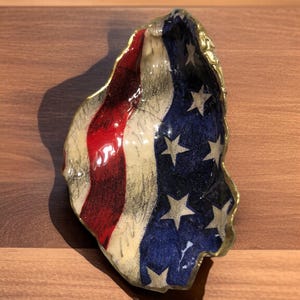 May include: A decorative oyster shell dish painted with the American flag design. The dish features red and white stripes, a blue field with white stars, and a gold-colored rim. The shell is placed on a wooden surface.