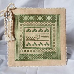 Puede incluir: Una pieza cuadrada de punto de cruz beige con un diseño geométrico verde. El diseño incluye un cuadrado central con patrones florales y geométricos. Una cuerda crema con borlas y un dije plateado cuelga de la esquina superior izquierda. Un artículo decorativo.